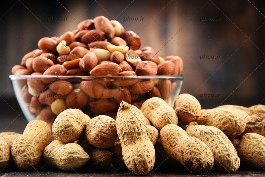 عکس بادام زمینی هایی با پوست روی زمین و بادام زمینی های پوست کنده داخل کاسه شیشه ای در پس زمینه چوبی
