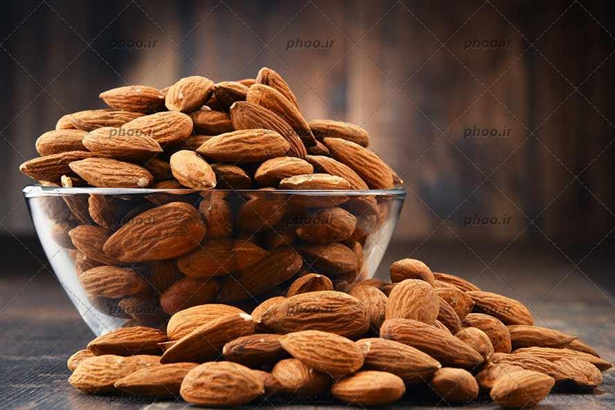 عکس بادام روی زمین و داخل کاسه شیشه ای در پس زمینه چوبی