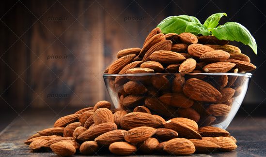 عکس بادام روی زمین و داخل کاسه شیشه ای با یک برگ ریحان در پس زمینه چوبی