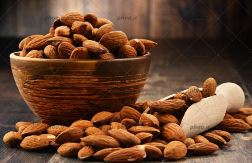 عکس بادام هایی در کاسه و قاشق چوبی و روی زمین در پس زمینه چوبی