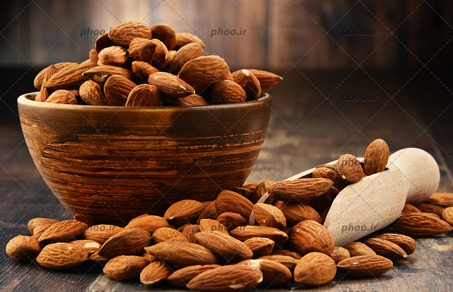 عکس بادام هایی در کاسه و قاشق چوبی و روی زمین در پس زمینه چوبی