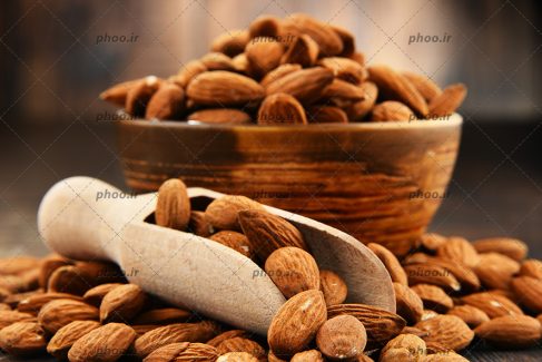 عکس بادام هایی در کاسه و قاشق چوبی و روی زمین در پس زمینه چوبی