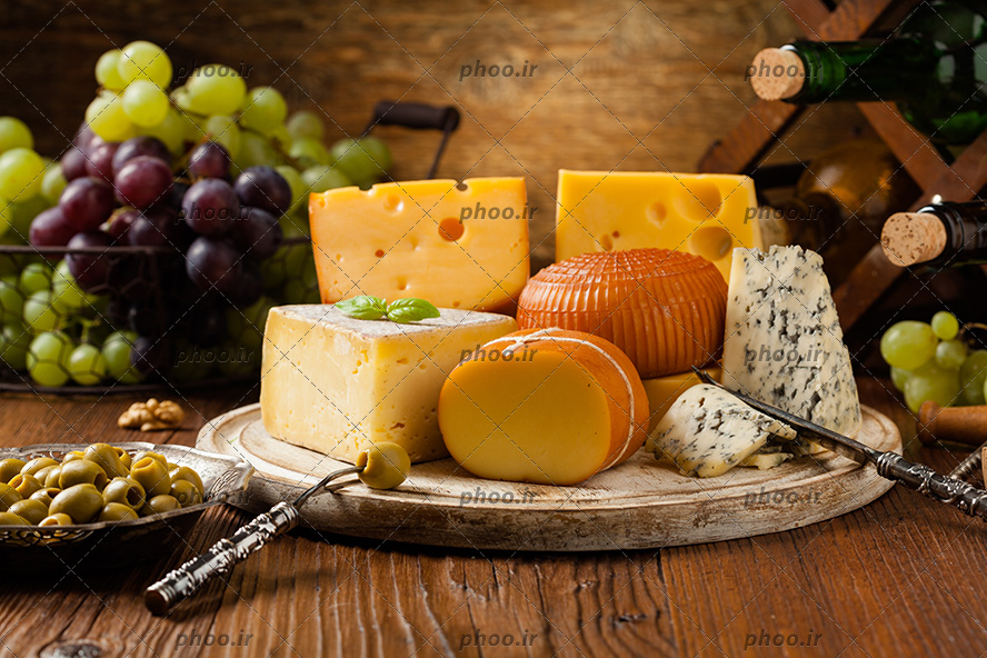 عکس انواع پنیر روی تخته چوبی در کنار انگور و زیتون و نوشیدنی در پس زمینه چوبی