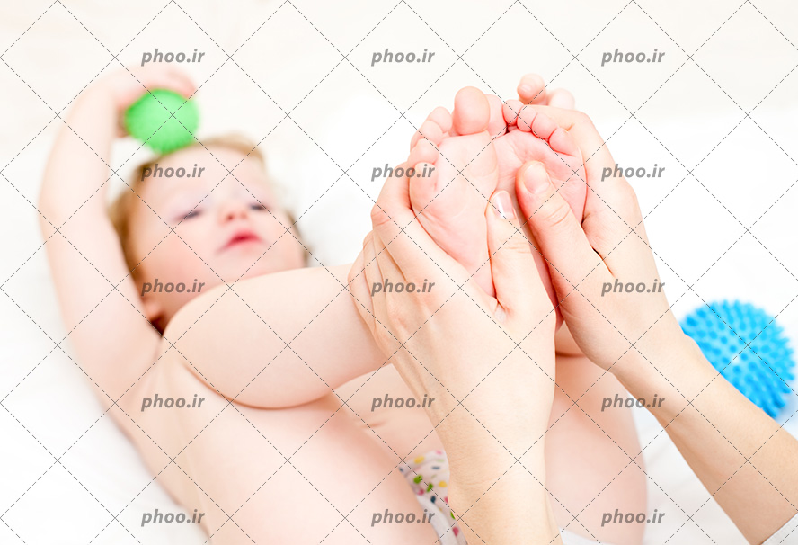 عکس با کیفیت کودک با یک توپ سبز در دست و مادر در حال ماساژ دادن پا های او