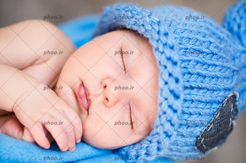 عکس با کیفیت نوزاد خوابیده با کلاه آبی بافتنی روی پتوی آبی