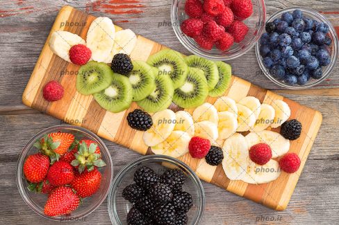 عکس میوه های برش خورده روی تخته چوبی و کاسه های شیشه ای از میوه
