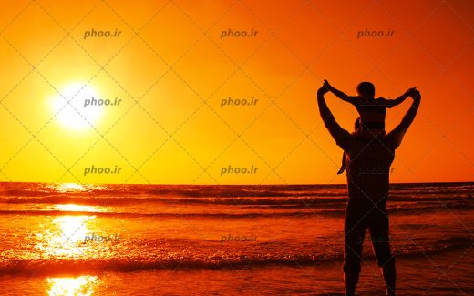 عکس با کیفیت پسر بچه بر روی دوش پدر در کنار دریا در مقابل غروب خورشید
