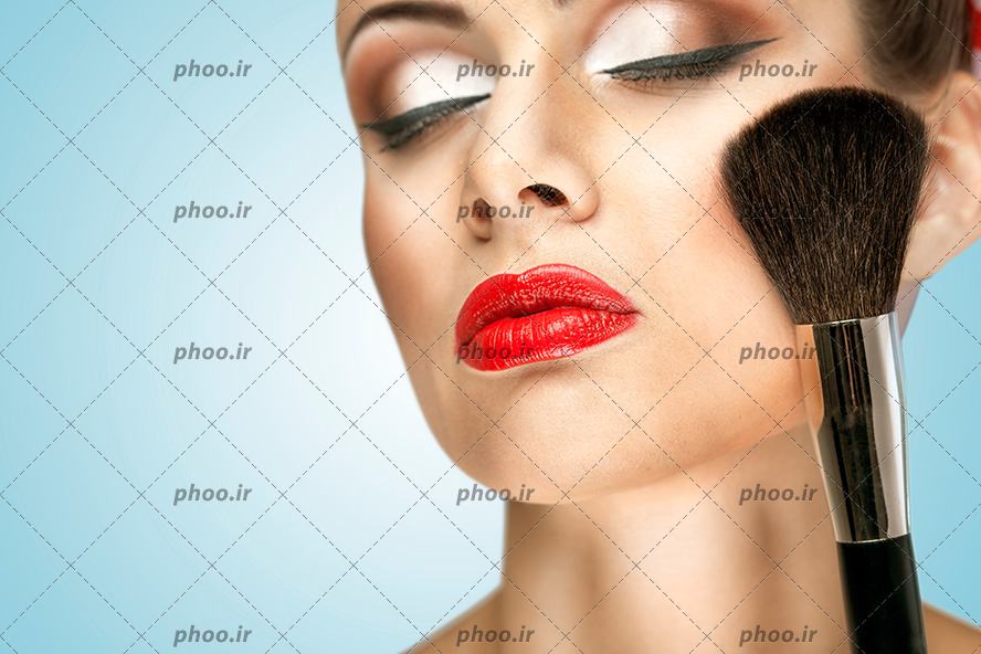 عکس خانم با چشم های بسته و آرایش صورت در حال رژ گونه زدن با براش آرایشی در پس زمینه آبی