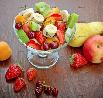 عکس میوه های تازه در ظرف شیشه ای و روی زمین در پس زمینه چوبی