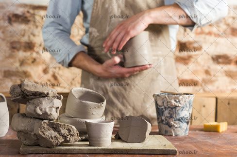عکس با کیفیت سفالگر ایستاده پشت میز و ظرف های سفالی و گِل روی میز