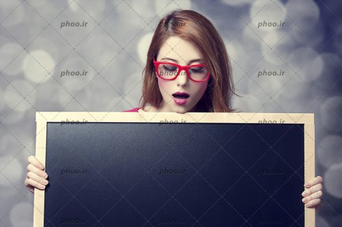 عکس با کیفیت خانم با عینک قاب قرمز و یک تخته سیاه با قاب چوبی در دست
