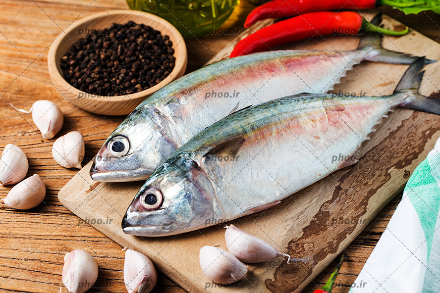 عکس دو ماهی خام در کنار ادویه و سیر و فلفل در پس زمینه چوبی