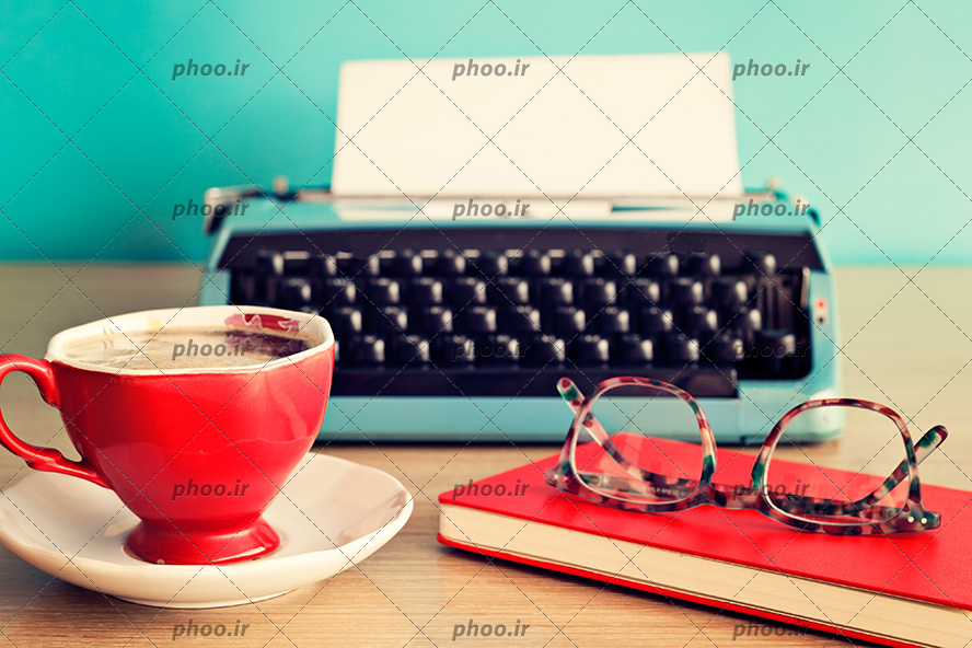 عکس دستگاه تایپ قدیمی در کنار فنجان قهوه و کتاب روی میز در پس زمینه آبی