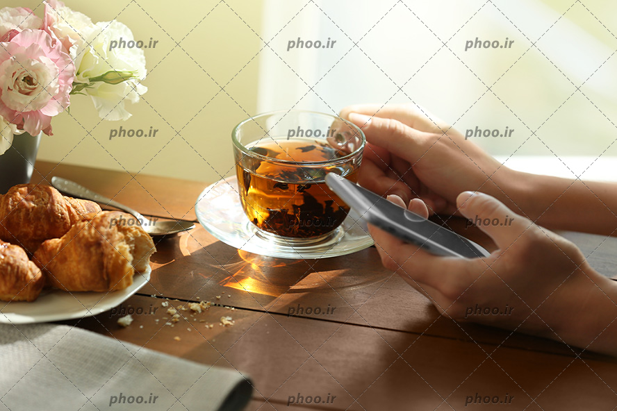 عکس شخصی که فنجان چای را در دست گرفته و به تلفن همراه نگاه می کند