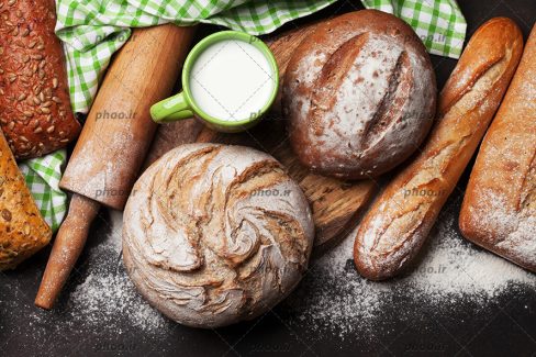 عکس نان ها و کیک های پخته شده با شکل های متفاوت در کنار یک لیوان شیر و وردنه و آرد در پس زمینه مشکی