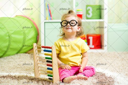 عکس کودک بامزه با مو های طلایی و عینک در کنار چرتکه در اتاق