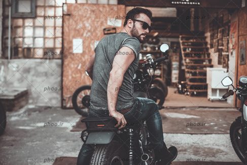 عکس مرد جوان با عینک آفتابی نشسته روی موتور سیکلت جلوی گاراژ یا تعمیرگاه