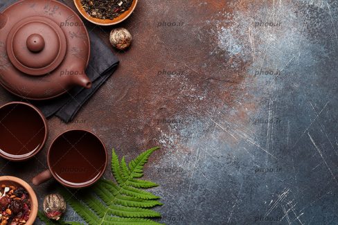 عکس دو فنجان چای در کنار قوری در پس زمینه قهوه ای