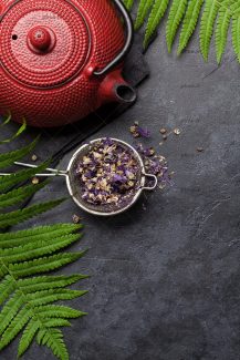 عکس قوری قرمز رنگ در کنار صافی با گل های بنفش روی میز سنگی