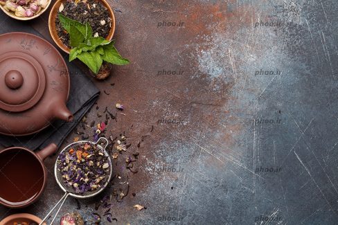 عکس یک فنجان چای در کنار قوری و گل بنفش و گل محمدی روی میز سنگی