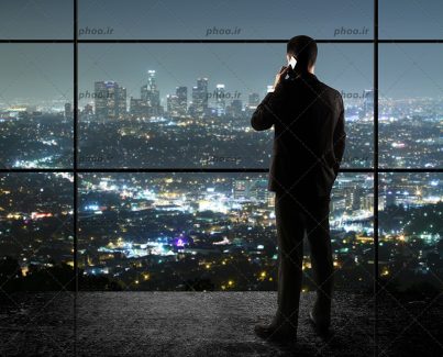 عکس تاجری که در دفتر شبانه با تلفن صحبت می کند و به شهر نگاه می کند