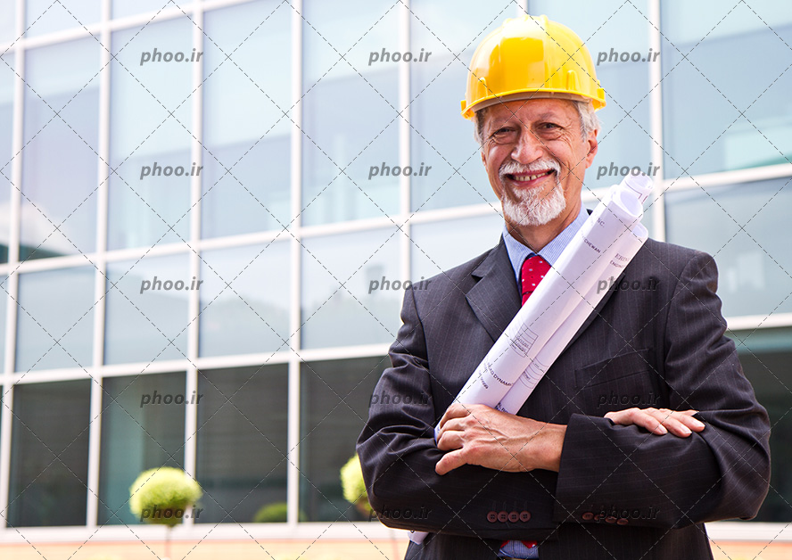 عکس مهندس با کلاه ایمنی و نقشه ساختمان در دست ایستاده در کنار ساختمان بلند