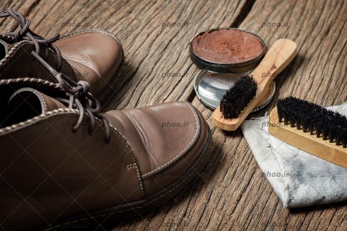 عکس کفش قهوه ای چرمی در کنار واکس قهوه ای و فرچه در پس زمینه چوبی