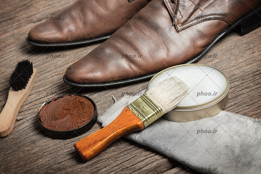 عکس کفش مردانه قهوه ای در کنار واکس قهوه ای و واکس براق کننده کفش در پس زمینه چوبی