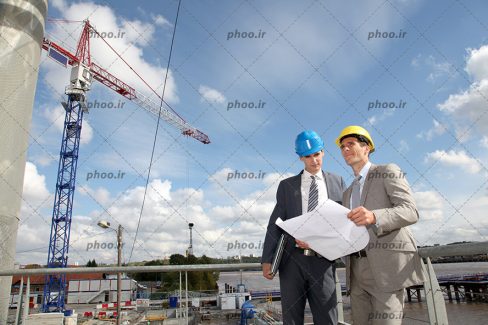 عکس دو مهندس با کت شلوار در حال بررسی نقشه ساختمان و مشورت کردن با هم