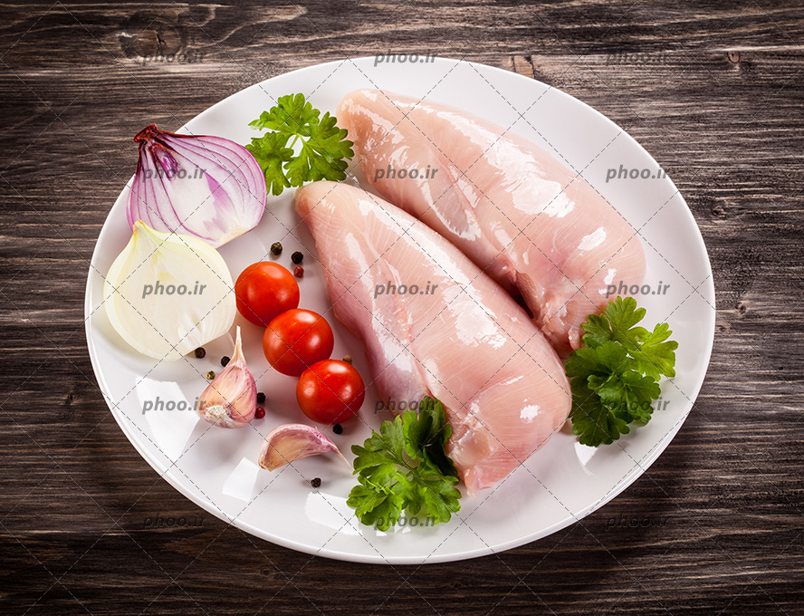 عکس فیله مرغ در کنار پیاز و گوجه و سیر در بشقاب سفید روی میز چوبی