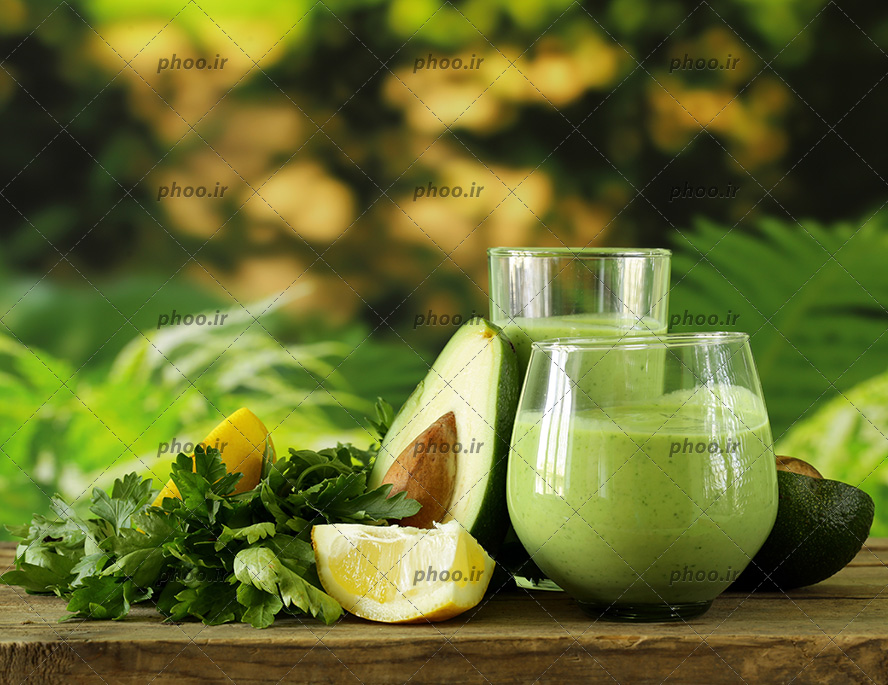 عکس آووکادو در کنار سبزی و لیمو و لیوان آبمیوه روی میز چوبی